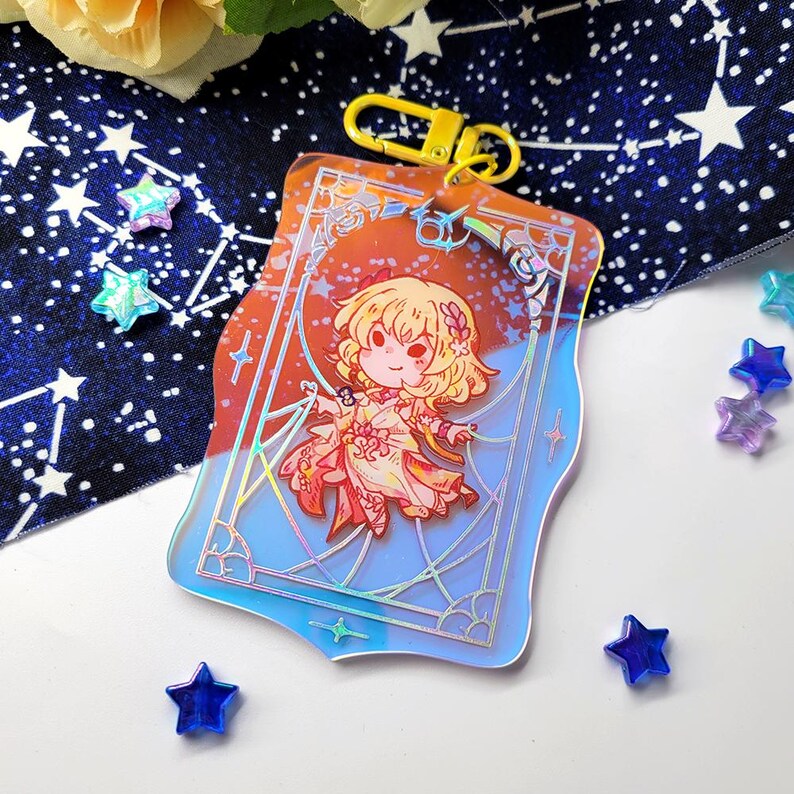 May include: A clear acrylic keychain featuring a cartoon character with blonde hair and a floral design. The keychain has a gold-colored clasp and a blue and orange gradient background. Decorative star-shaped beads are scattered around the keychain.