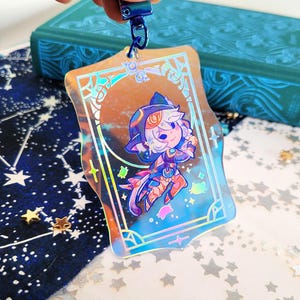 May include: Holographic acrylic charm featuring a cartoon character with a cat-like appearance, wearing a blue and orange outfit. The charm has a blue metal clasp and a decorative border. The background includes a starry pattern and a teal book.