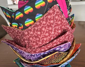 May include: A stack of handmade bowl cozies in various colorful patterns. The cozies feature designs like rainbow hearts, lions, and floral prints. The edges are trimmed with contrasting colors, and the cozies are stacked on a wooden surface.