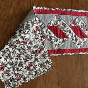 May include: A quilted Christmas table runner with a festive design. The runner features a pattern of red, white, and gray geometric shapes and a section with a repeating pattern of ornaments, holly, and berries. The runner is displayed on a wooden table.