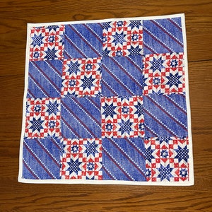 May include: A square, patriotic-themed quilt with a repeating pattern of red, white, and blue stars and stripes. The quilt is bordered with a white edge and is set on a wooden surface.
