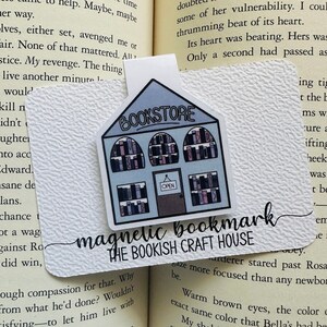 Peut inclure: Marque-page magnétique blanc avec un design de librairie. Le marque-page présente un bâtiment bleu avec le mot "BOOKSTORE" et un panneau "OPEN". Le texte "magnetic bookmark THE BOOKISH CRAFT HOUSE" est également visible.