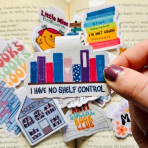 May include: A white magnetic bookmark featuring a colourful illustration of a bookshelf filled with books. The text "I Have No Shelf Control" is printed below the bookshelf. The bookmark is surrounded by other colourful book-themed stickers.