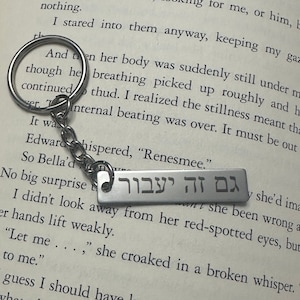 Peut inclure: Un porte-clés argenté avec une étiquette rectangulaire sur laquelle est inscrit "גם זה יעבור" en hébreu. Le porte-clés est posé sur un livre avec du texte.