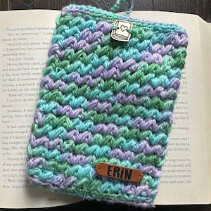 May include: A handmade crocheted book sleeve in blue, green, and lilac hues. It has a wooden heart button and a leather tag with the name "ERIN". Designed to protect books, it measures approximately 20 cm in length.