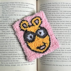 May include: A handmade, square, pink, tufted coaster featuring a cartoon character with yellow fur, black eyes, and a smiling face. The coaster is placed on an open book.
