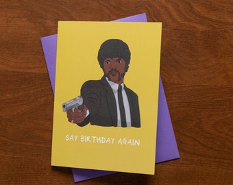 May include: A yellow greeting card with a cartoon illustration of a man in a black suit holding a gun. The text on the card says "Say Birthday Again".