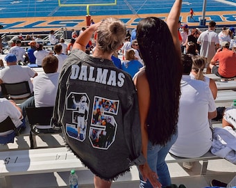 May include: Two people wearing denim jackets are sitting in a stadium. The person in the foreground is wearing a jacket with the name "DALMAS" and the number "35" on the back. The jacket has a photo of a football player on the back. The person in the background is wearing a denim jacket with long black hair.