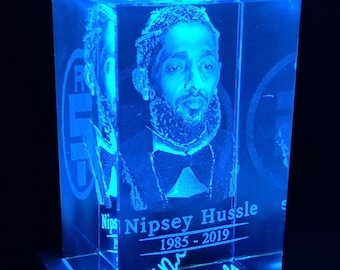 Könnte beinhalten: Ein blauer, beleuchteter Kristallwürfel mit einem 3D-Bild des Rappers Nipsey Hussle. Der Würfel trägt den Text "Nipsey Hussle 1985 - 2019" und eine Unterschrift auf der Unterseite.