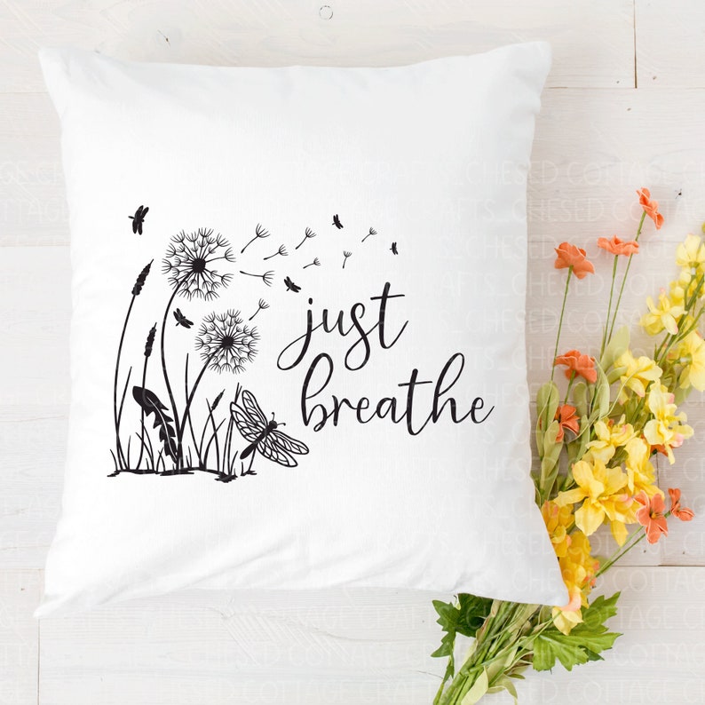 May include: White square cushion featuring a black illustration of dandelions, grass, and a dragonfly. The words "just breathe" are written in a cursive font. Orange and yellow flowers are in the background.
