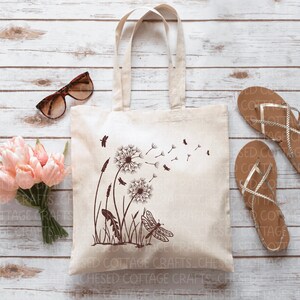 May include: A beige canvas tote bag featuring a brown dandelion and dragonfly design. The bag has two handles and is placed on a light wood surface. Other items include sunglasses, pink flowers, and sandals.