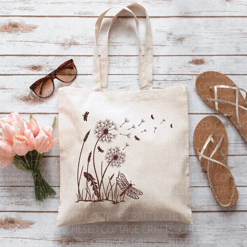 May include: A beige canvas tote bag featuring a brown dandelion and dragonfly design. The bag has two handles and is placed on a light wood surface. Other items include sunglasses, pink flowers, and sandals.