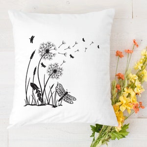 May include: White square cushion featuring a black dandelion and dragonfly design. The design includes dandelions with seeds blowing, grass, and a dragonfly. The cushion is on a white wooden surface with flowers.