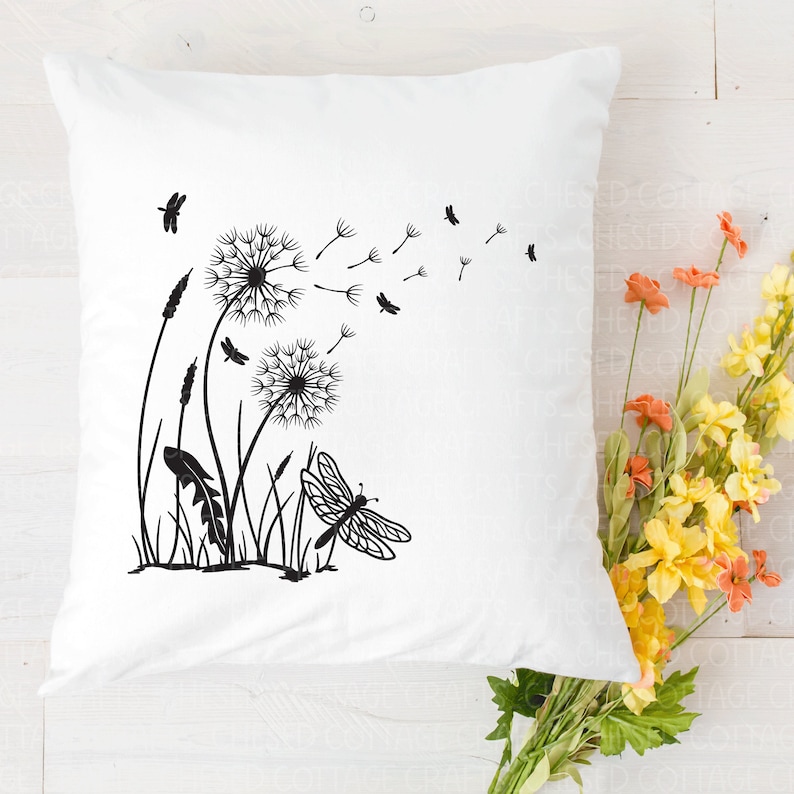 May include: White square cushion featuring a black dandelion and dragonfly design. The design includes dandelions with seeds blowing, grass, and a dragonfly. The cushion is on a white wooden surface with flowers.
