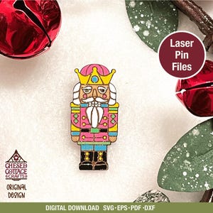 May include: A colorful wooden nutcracker pin with a pink jacket, blue pants, and a gold crown. The pin is surrounded by red bells and green leaves. The text "Laser Pin Files" is in a maroon circle.