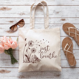 May include: A cream tote bag featuring a brown dandelion design and the words "just breathe." The bag is placed next to sunglasses, pink tulips, and brown sandals. The bag has two handles.