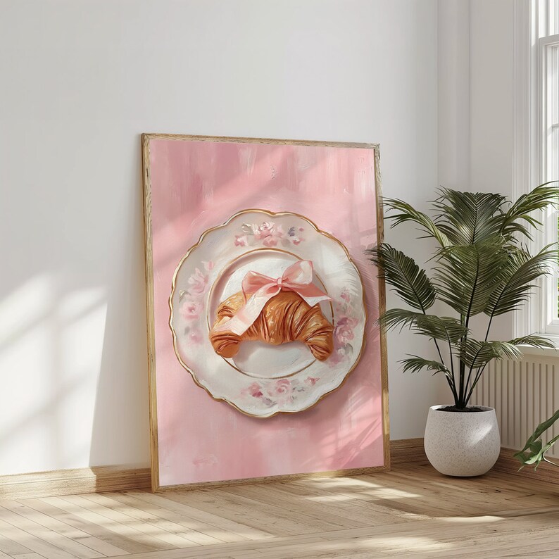 May include: A framed artwork featuring a croissant on a decorative plate with a pink bow. The plate has a gold rim and floral accents, set against a pink background. The artwork is displayed in a room with a plant.