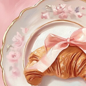 May include: A still-life painting of a croissant tied with a pink bow, resting on a white plate with a gold rim and floral accents. The background is a soft pink, creating a delicate and inviting aesthetic.