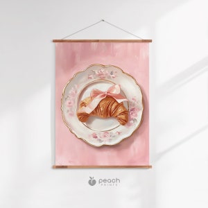 May include: A pink wall hanging featuring a still life of a croissant on a decorative plate. The plate has a gold rim and floral designs. A pink bow adorns the croissant. The background is a solid pink color. The print is held by wooden bars.