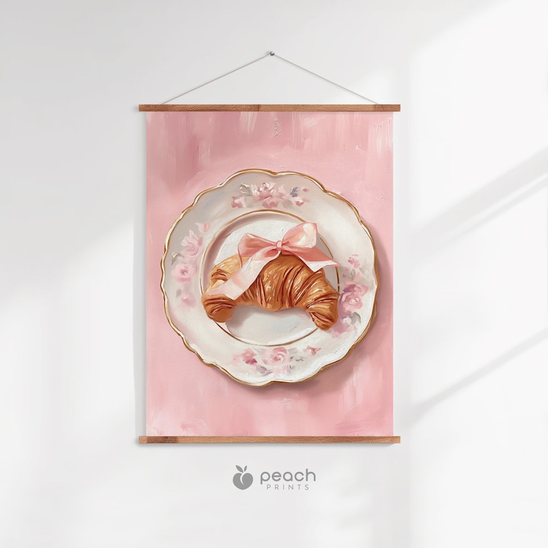 May include: A pink wall hanging featuring a still life of a croissant on a decorative plate. The plate has a gold rim and floral designs. A pink bow adorns the croissant. The background is a solid pink color. The print is held by wooden bars.