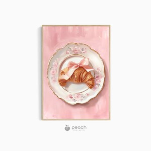 May include: A framed print featuring a croissant on a decorative plate with a pink bow. The plate has a floral design and gold trim, set against a pink background. The print has a wooden frame and the Peach Prints logo.