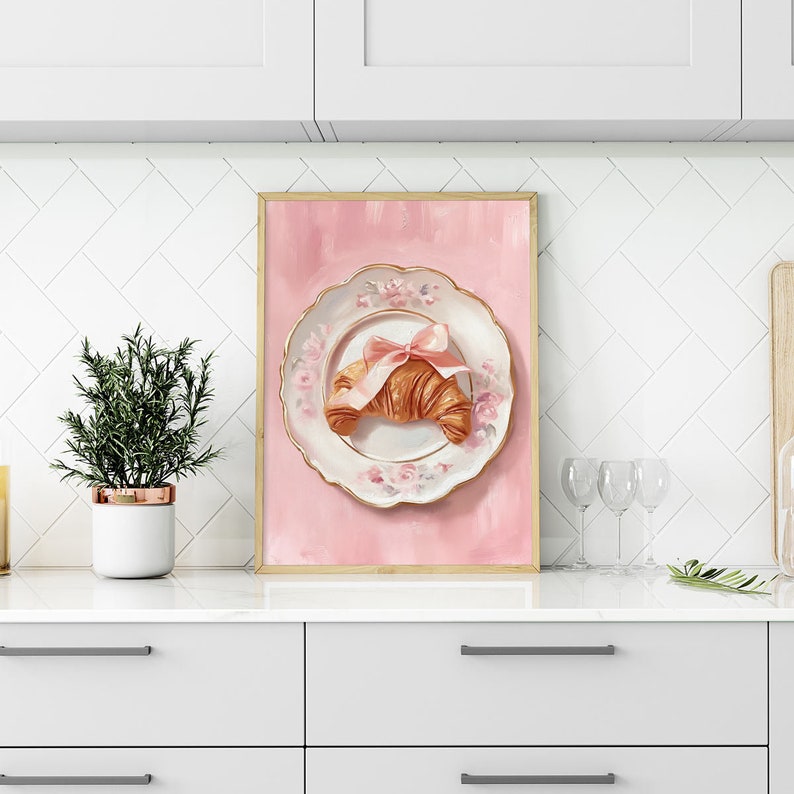 May include: A framed print featuring a croissant tied with a pink bow on a decorative plate with floral accents. The plate is set against a pink background. The print has a gold frame and is displayed on a white countertop.