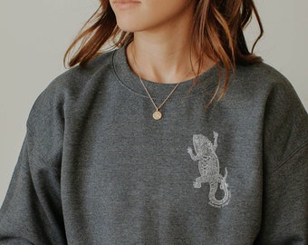 May include: Dark grey crew neck sweatshirt featuring a lizard design on the upper left chest. The sweatshirt is worn with blue jeans. A person is holding a white mug.