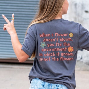 May include: A dark gray t-shirt with the text "PARA professional" on the front and the text "When a flower doesn't bloom, you fix the environment in which it grows, not the flower." on the back. The text is in a pink color with a flower graphic.