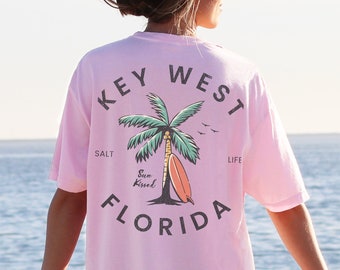 Puede incluir: Camiseta rosa claro con un gráfico en la espalda que dice "Key West Florida" con un diseño de palmera y tabla de surf. La camiseta es de gran tamaño y la lleva una persona de pie cerca del mar.