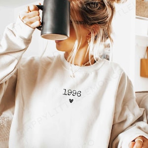 May include: Sand-colored sweatshirt with the year "1996" and a small black heart printed on the front. The sweatshirt has a crew neck and long sleeves. A person is holding a black mug.