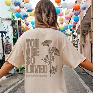 May include: A beige t-shirt with the words "YOU ARE SO LOVED" printed on the back, along with a floral design and a Bible verse. The shirt is a relaxed fit. The image is taken outdoors with colorful paper lanterns in the background.