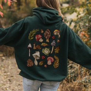 May include: A dark green hooded sweatshirt with a colorful mushroom and frog design on the back. The design features various mushrooms, a frog, a snail, leaves, and pine cones.