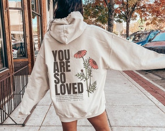 Puede incluir: Una sudadera con capucha de color crema con el texto "You are so loved" y un diseño floral en la espalda. La sudadera es de gran tamaño y tiene una capucha con cordón.