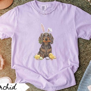 May include: Orchid-colored t-shirt featuring an Easter-themed graphic. The design includes a dachshund wearing bunny ears and holding a carrot, with chicks and eggs. The shirt has a distressed neckline and the word "Orchid" on a white banner.