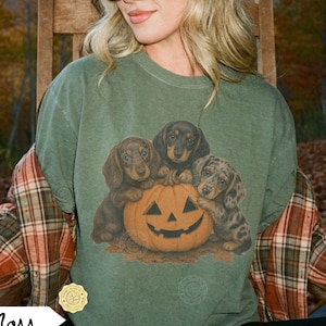 May include: A moss green short-sleeve t-shirt featuring a graphic print of three dachshund puppies huddled around a carved orange jack-o'-lantern pumpkin. The design is centered on the chest, showcasing the dogs with brown and dapple fur.