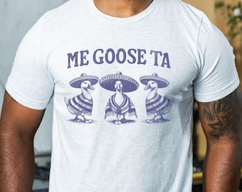 May include: A white t-shirt with a purple graphic of three geese wearing sombreros and ponchos. The text "ME GOOSE TA" is printed above the geese.