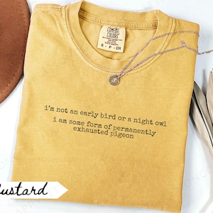 May include: Mustard-colored Comfort Colors t-shirt with the text "i'm not an early bird or a night owl i am some form of permanently exhausted pigeon." A silver necklace with a pendant is on the shirt.
