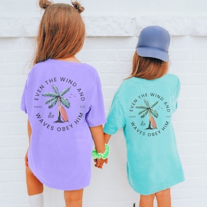 May include: Two children wearing purple and turquoise t-shirts with a palm tree and surfboard graphic. The text on the shirts reads "Even the wind and waves obey him" with the scripture reference "Mark 4:41".