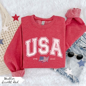 May include: A red heather sweatshirt with the word "USA" in a white, collegiate-style font. Below the text is a graphic of the American flag and the years 1776 and 2026. The sweatshirt is paired with denim shorts and sunglasses.