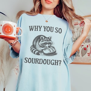 May include: A light blue t-shirt with a raccoon graphic and the text "WHY YOU SO SOURDOUGH?"