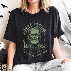 May include: Black t-shirt featuring a green graphic of Frankenstein's monster. The graphic reads "Just Trying To Keep It Together".