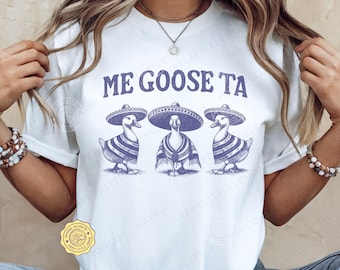 May include: A white short-sleeved t-shirt featuring a graphic print of three geese wearing sombreros and striped ponchos. Above the geese, the text reads ME GOOSE TA in a dark blue, distressed font. The shirt has a crew neckline and a relaxed fit.