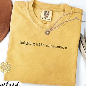 May include: Mustard-colored t-shirt with the text "mahjong with mahsisters" in black. The shirt has a crew neck and short sleeves. A silver necklace with a small pendant is on the shirt. The shirt is labeled "Comfort Colors".