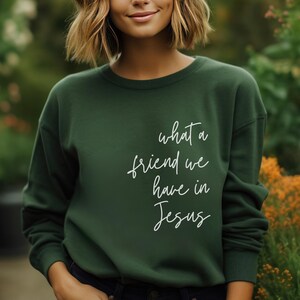 May include: A dark forest green crewneck sweatshirt featuring the white cursive text what a friend we have in Jesus printed on the front. The garment has long sleeves with ribbed cuffs and a ribbed neckline, providing a simple and clean design.