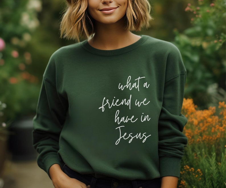 May include: A dark forest green crewneck sweatshirt featuring the white cursive text what a friend we have in Jesus printed on the front. The garment has long sleeves with ribbed cuffs and a ribbed neckline, providing a simple and clean design.