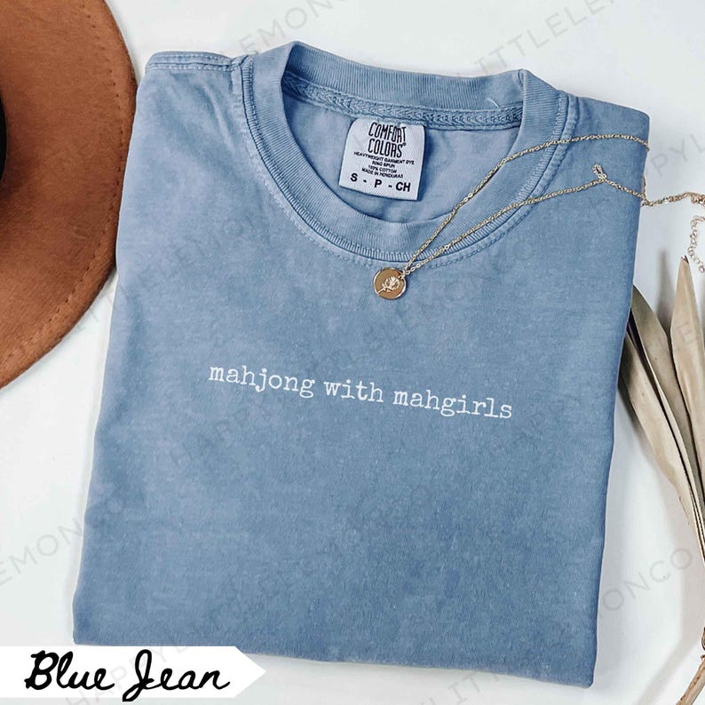 May include: A blue jean coloured t-shirt with the text "mahjong with mahgirls" in white. The shirt has a crew neck and short sleeves. A gold necklace with a pendant is draped across the shirt. The shirt is size S-CH.