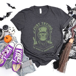 May include: A dark gray t-shirt with a green graphic of Frankenstein's monster. The graphic says "Just Trying To Keep It Together".