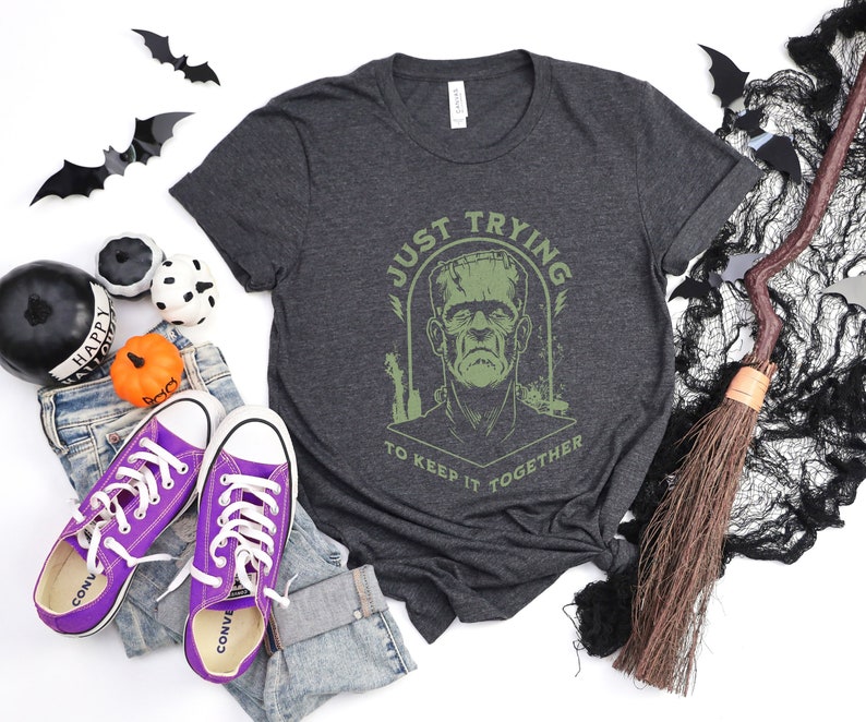 May include: A dark gray t-shirt with a green graphic of Frankenstein's monster. The graphic says "Just Trying To Keep It Together".
