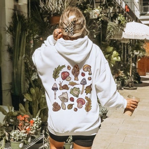 May include: White hoodie with a colorful mushroom and nature-themed graphic print on the back. The graphic includes mushrooms, leaves, a snail, a frog, and ladybugs.