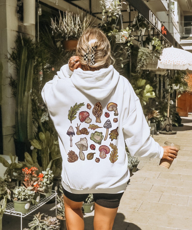 May include: White hoodie with a colorful mushroom and nature-themed graphic print on the back. The graphic includes mushrooms, leaves, a snail, a frog, and ladybugs.